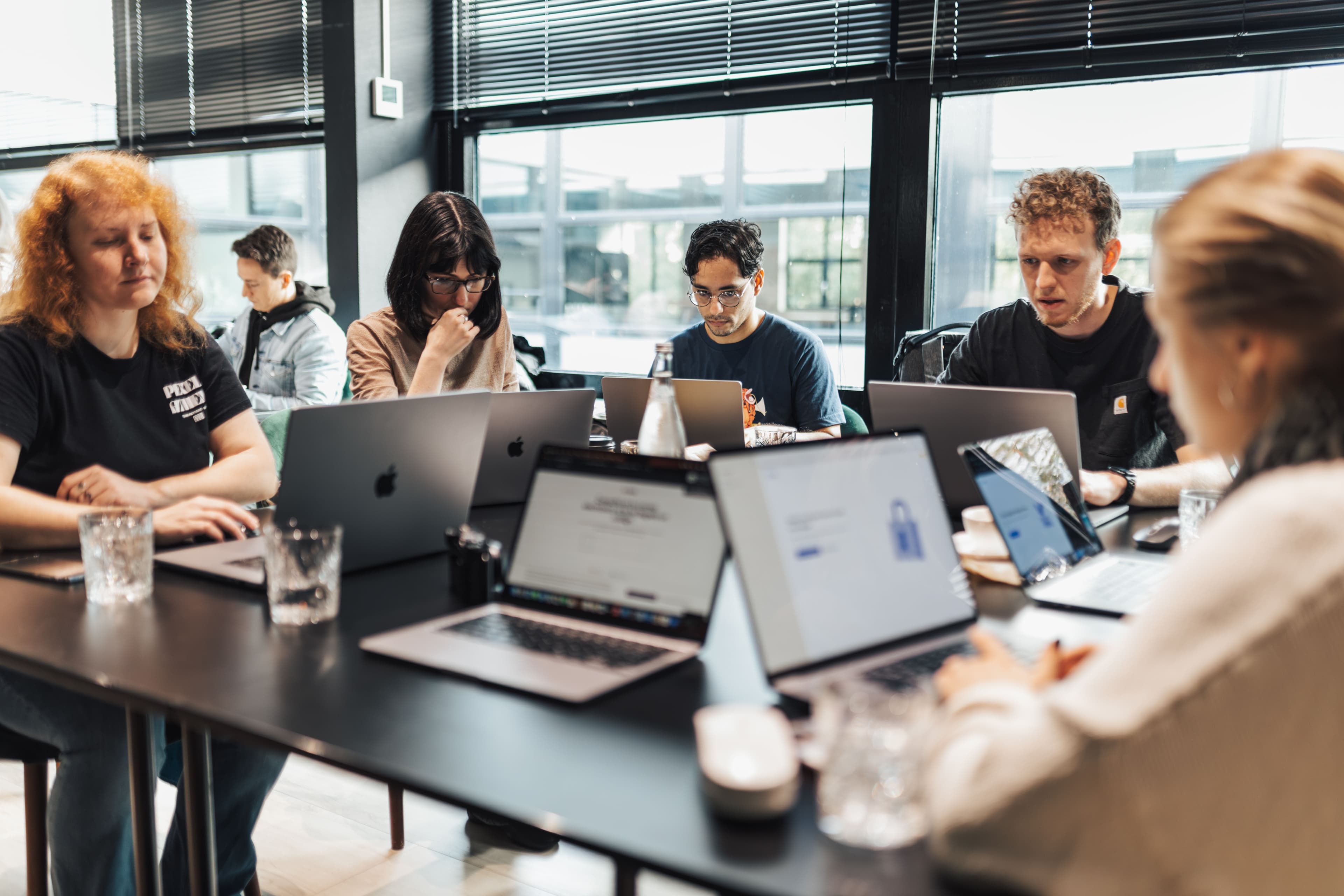 Deelnemers aan een workshop werken aan een lange tafel ieder op hun eigen laptop.